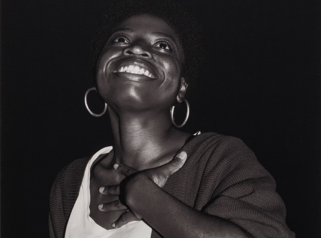 A monochrome photo of a person looking up, smiling, their hand on their neck. They are wearing a dark long-sleeved sweater over a white shirt.