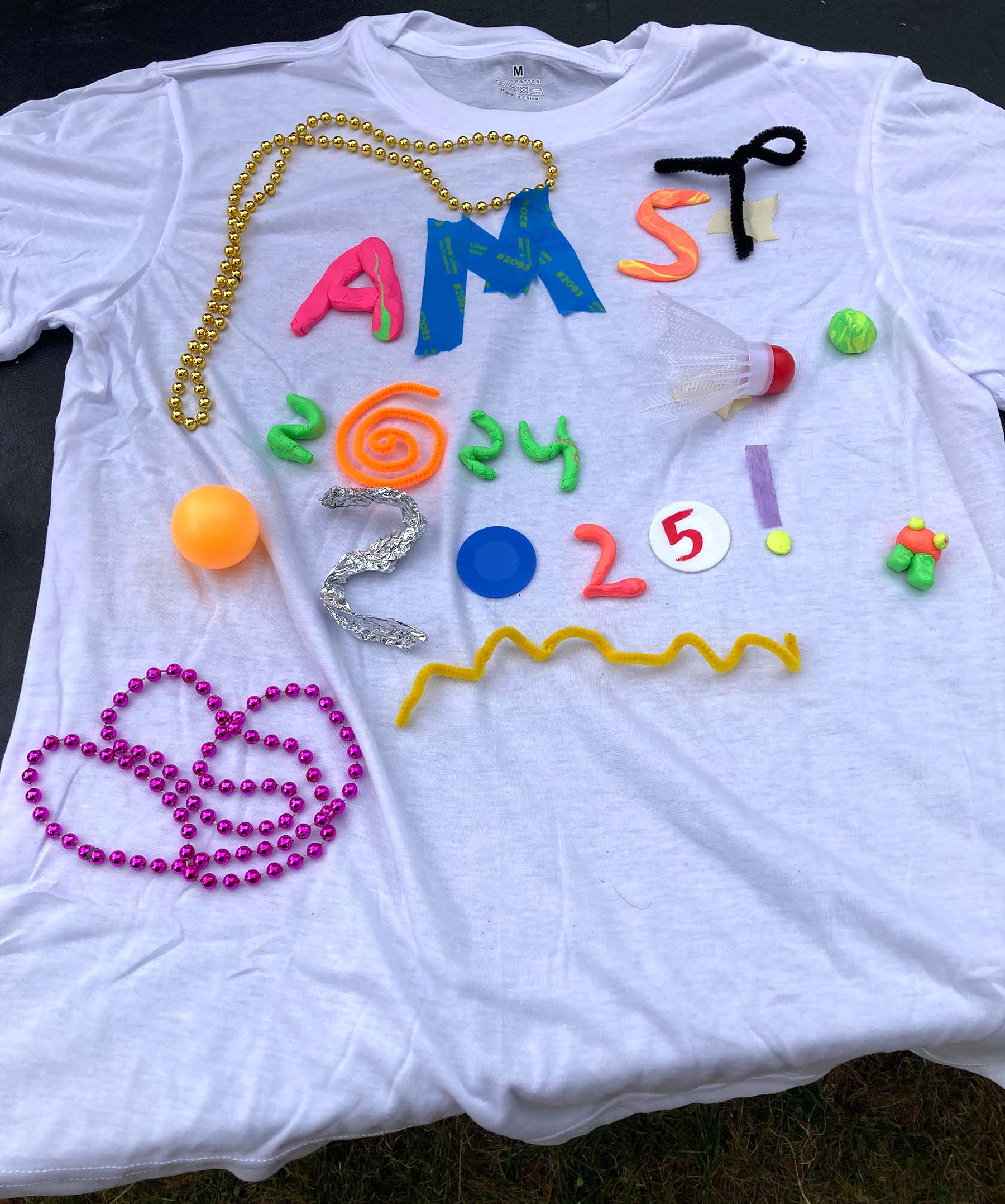 Whtie t-shirt decorated with beads, material, foil, pipe clearners, badminton birdie and other items spelling out AMST 2024–2025.