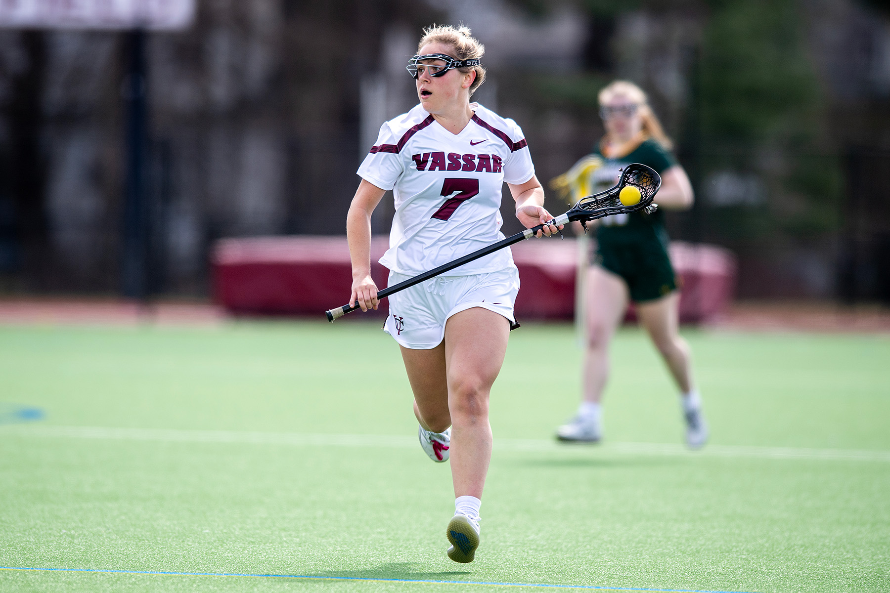 A lacrosse player running across a field in a white uniform, lacrosse stick, and wearing a facemask.