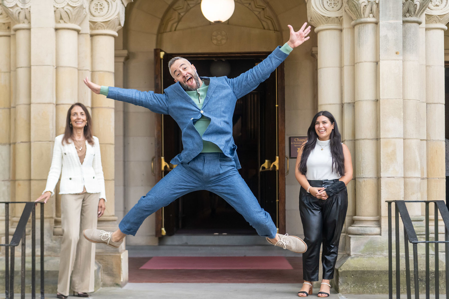 Person in a blue suit and green shirt jumping into the air with arms and legs outstretched while standing in an building entrance with a person on each side looking on.