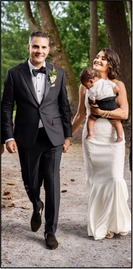 Two people walking dressed for a wedding in tux and dress. One person is carrying a young child while holding the other's hand.