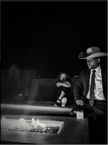 a dark photo of a person in suit and tie with a cowboy hat looking into an outside fire.