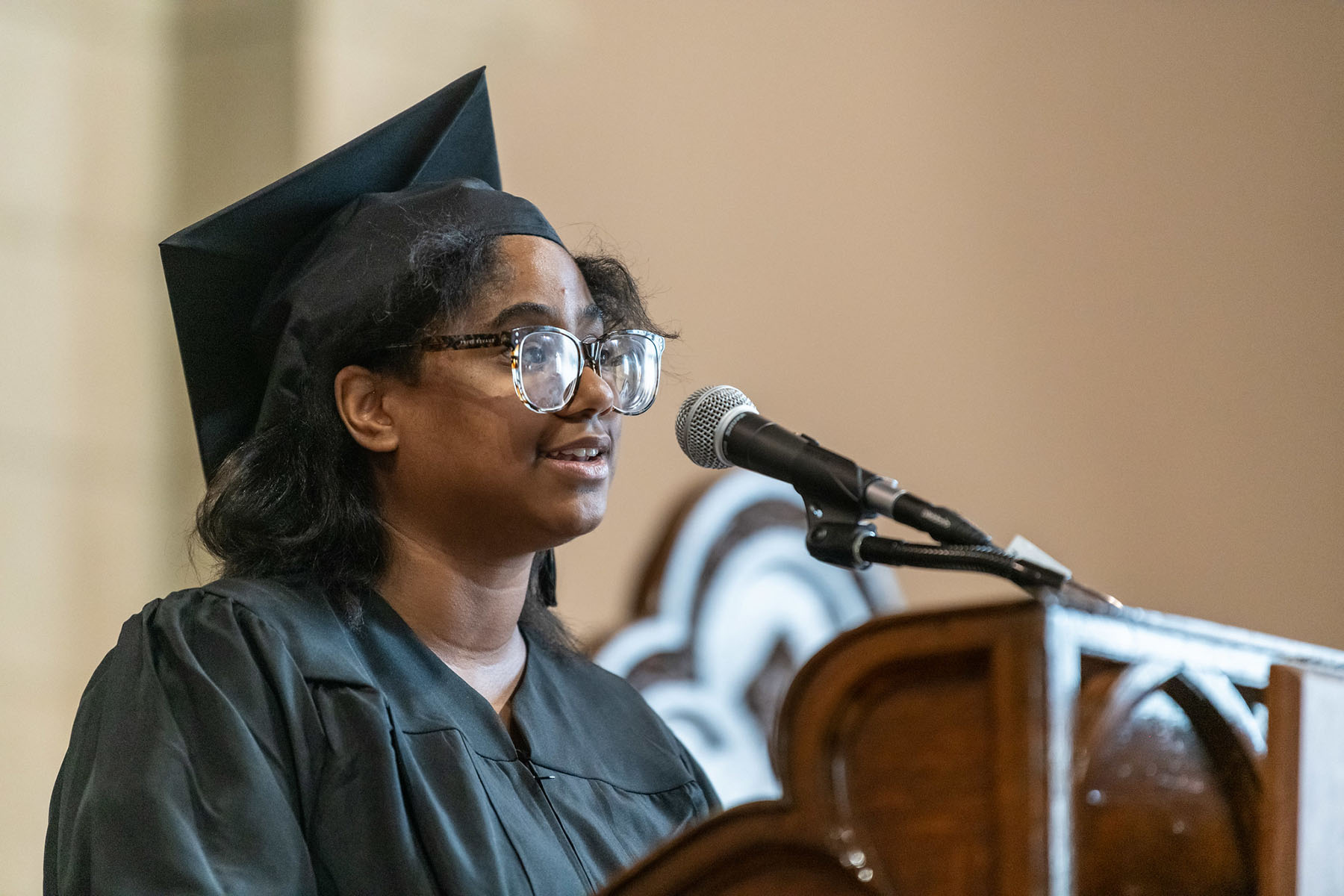 A person in a graduation cap and gown standing at a podium speaking. to an audience.