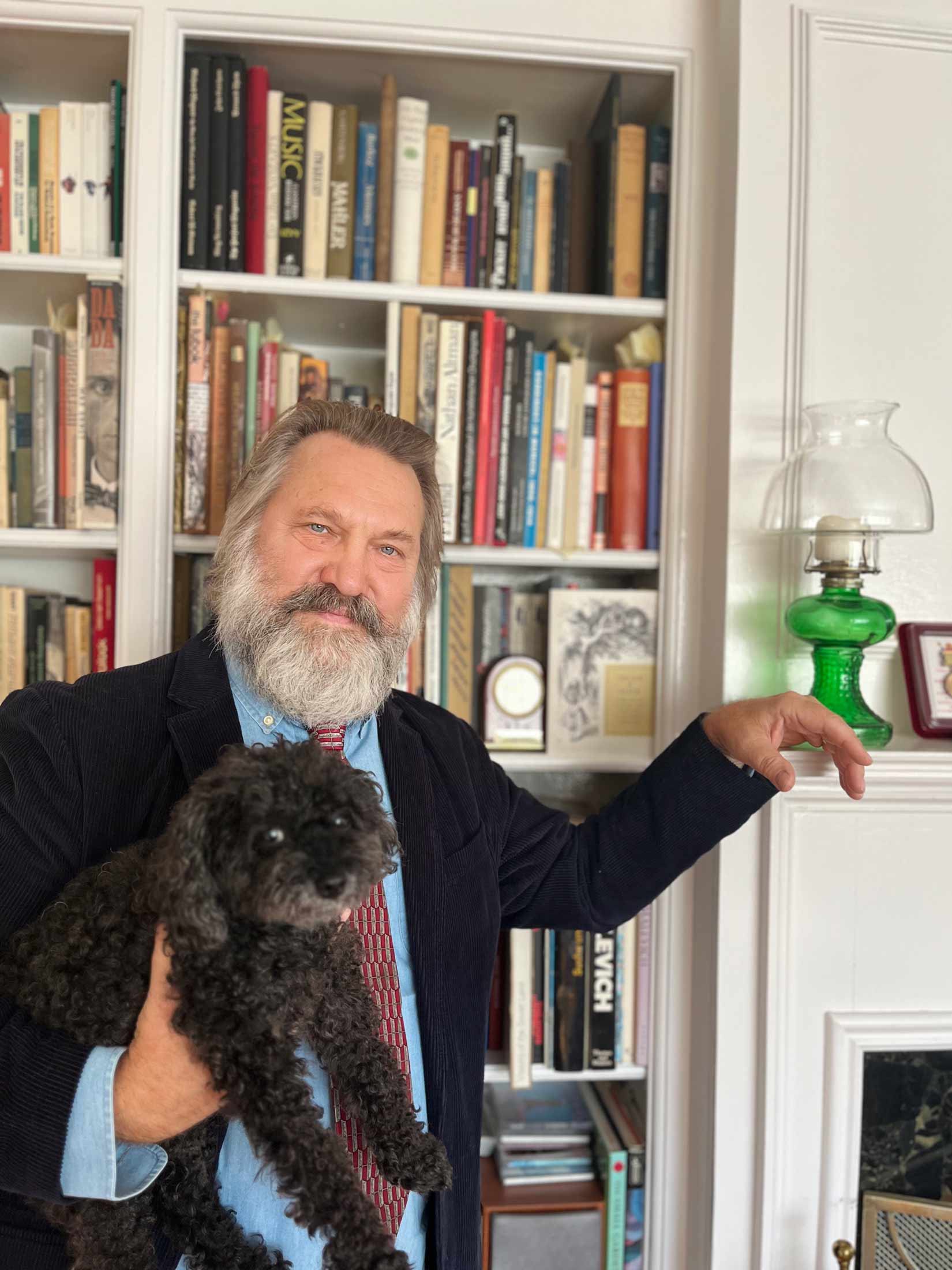 A photo of Nikolai Firtich holding his dog, standing in front of a bookshelf.