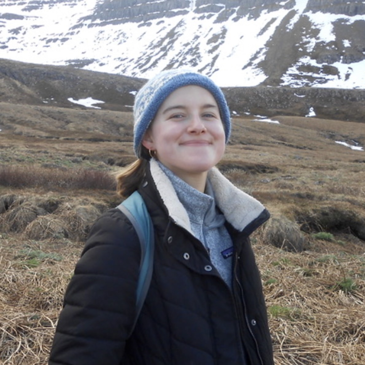 A headshot of Lily Shimpach, a person with long brown hair, a blue knitted hat, and a black jacket.