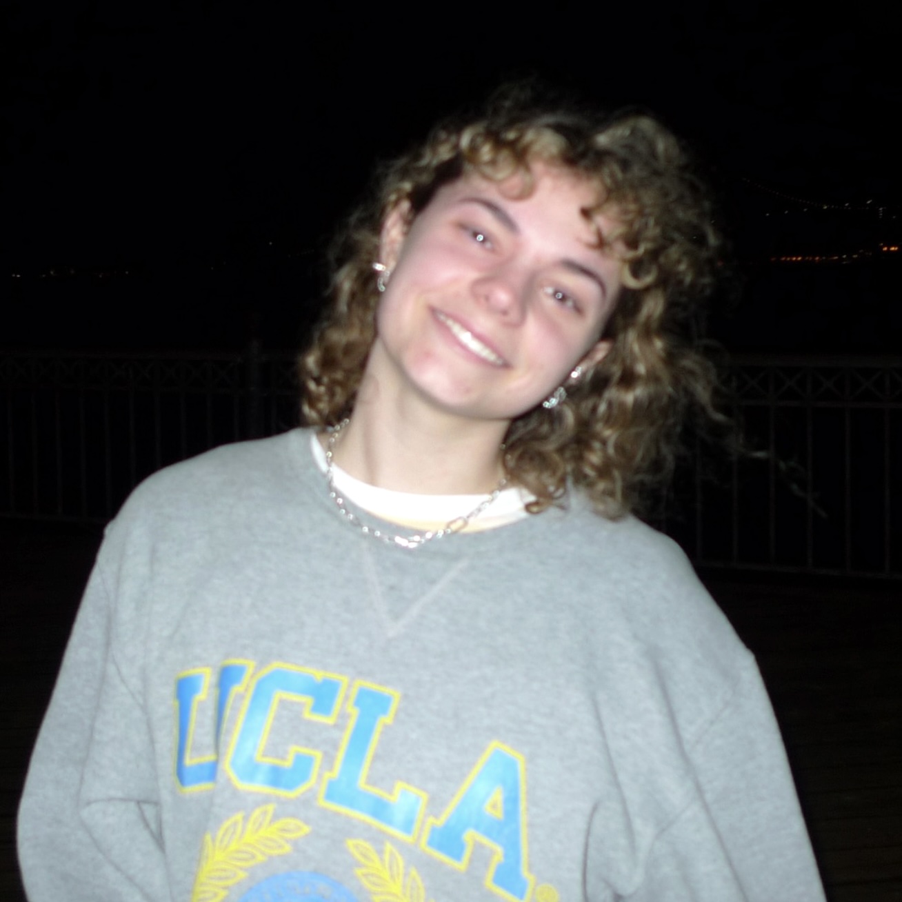 A headshot of Maisy Richardson, a person with long blond curly hair and a gray sweater with "UCLA" printed on it.