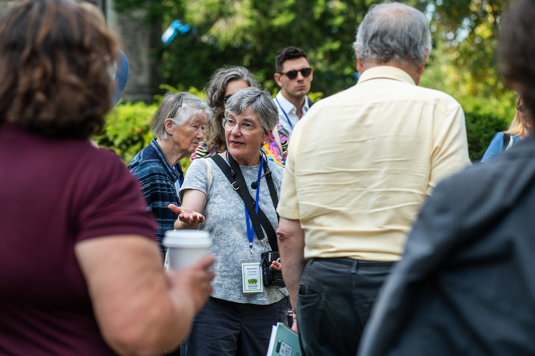 Person standing in the middle of a group of people speaking while outside.