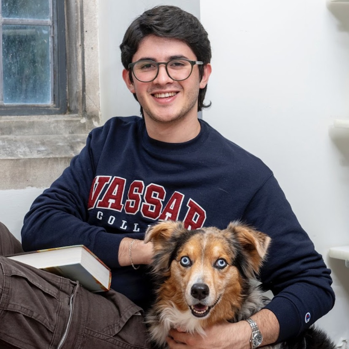 A headshot of Nico Reyes Cardozo, a person with dark brown hair, glasses, and a dark blue shirt with "Vassar College" printed on it.