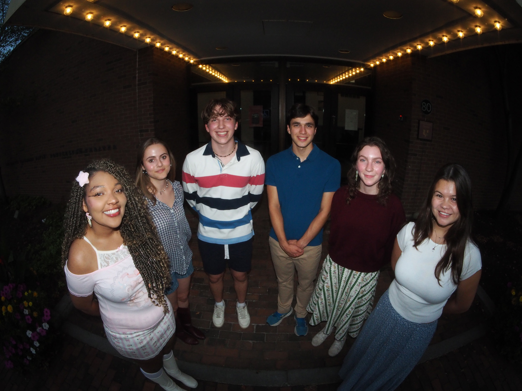 Six people standing in a half-circle on a dark stage with lights overhead.