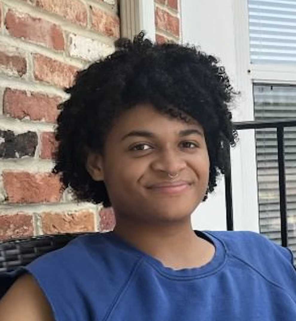 A portrait of Rae-Aila Crumble, a person with long black curly hair and a blue shirt.