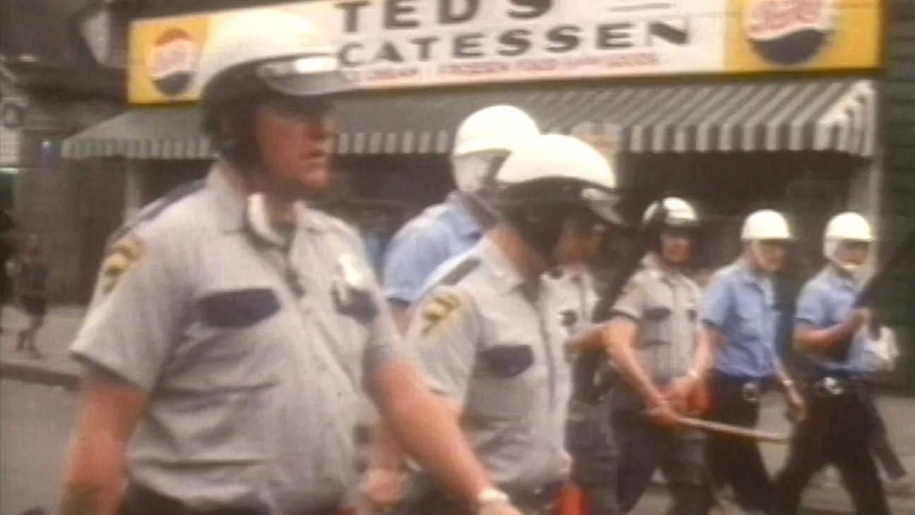 A blurry frame from the film "Power" by Yance Ford. It shows a group of police officers in helmets, holding batons and marching in a line.