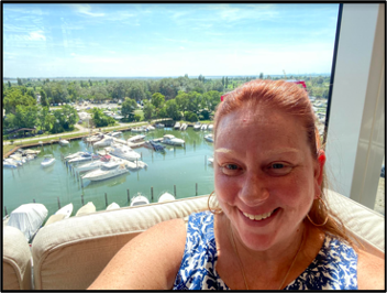 Person taking a selfie in a whicker chair with boats docked at a marina in the background.