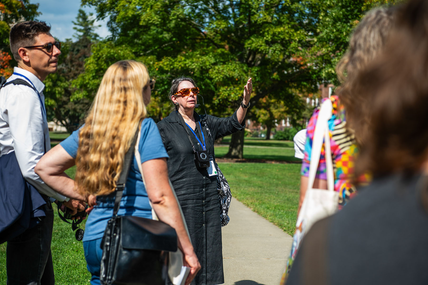 Person with rose colored glasses standing outside in the Vassar Quad speaking with their hand raise while giving a tour with others looking on.