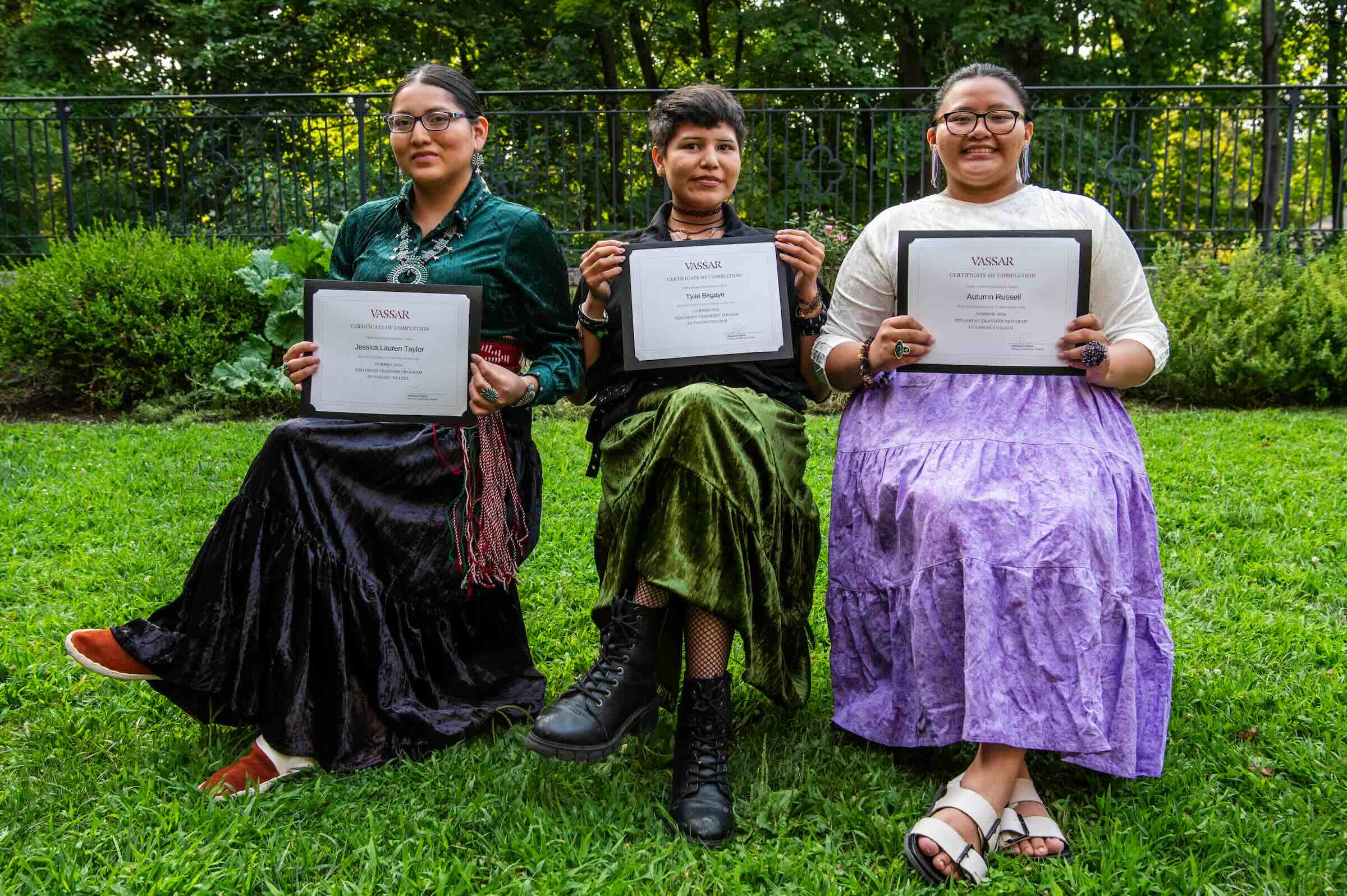 Three people seated in chairs on a lawn holding up diplomas and smiling.