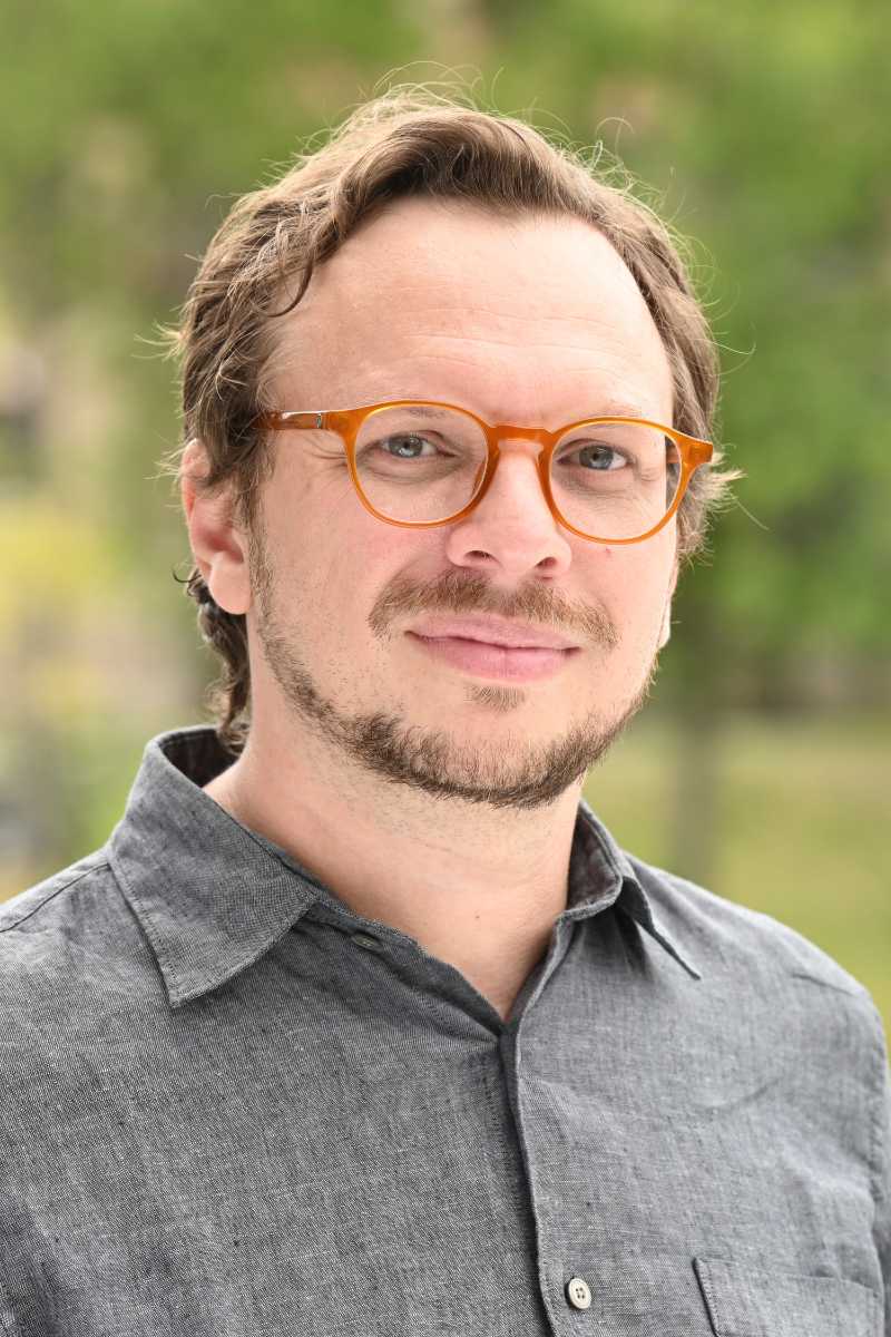 A portrait of Fabio Andrade, a person with short blond hair and beard, glasses, and a gray shirt.