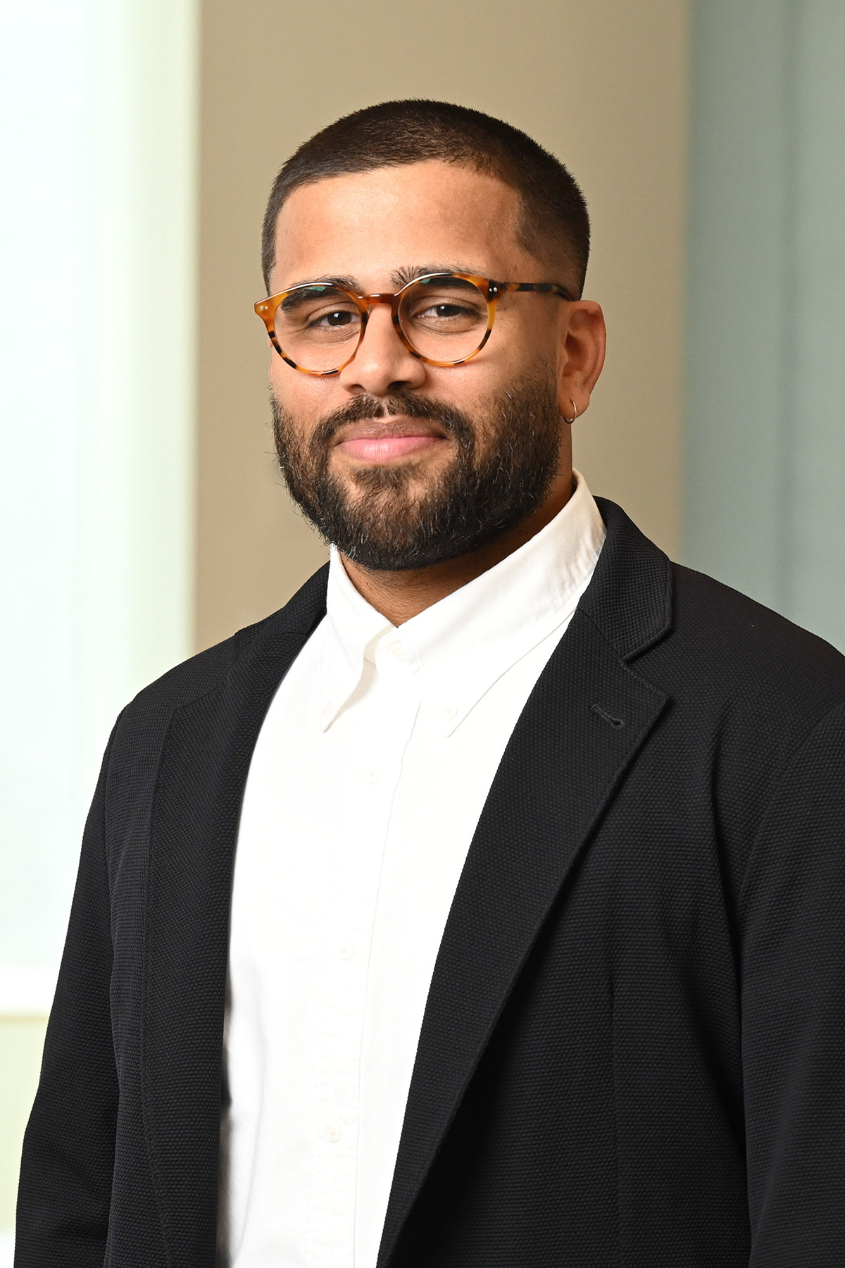 Portrait of a person in a bright, well-lit room wearing a long black suit-jacket over a buttoned-down white shirt. The person has glasses, dark hair and close-cropped mustache and beard.