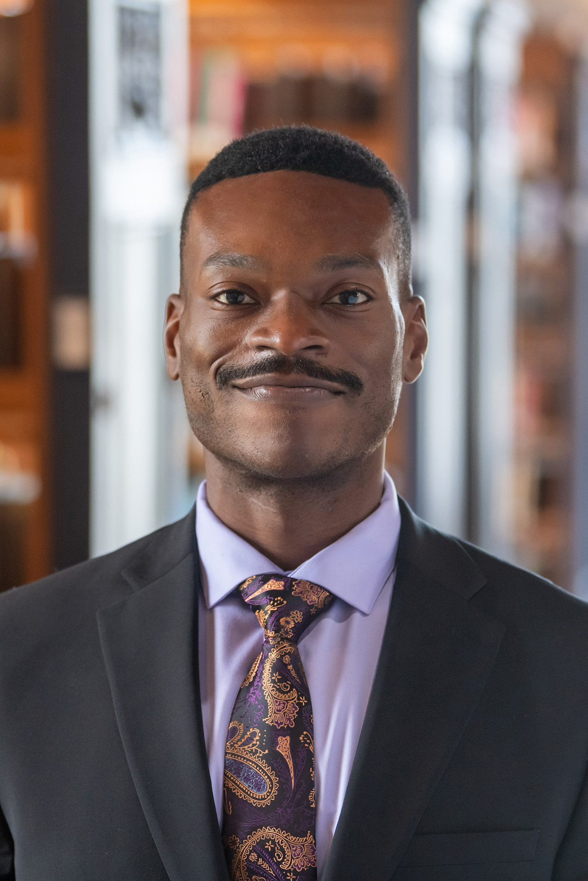 A person standing in a library with book shelves in the background wearing a suit and tie. The person has short cropped black hair and a tight black mustache.