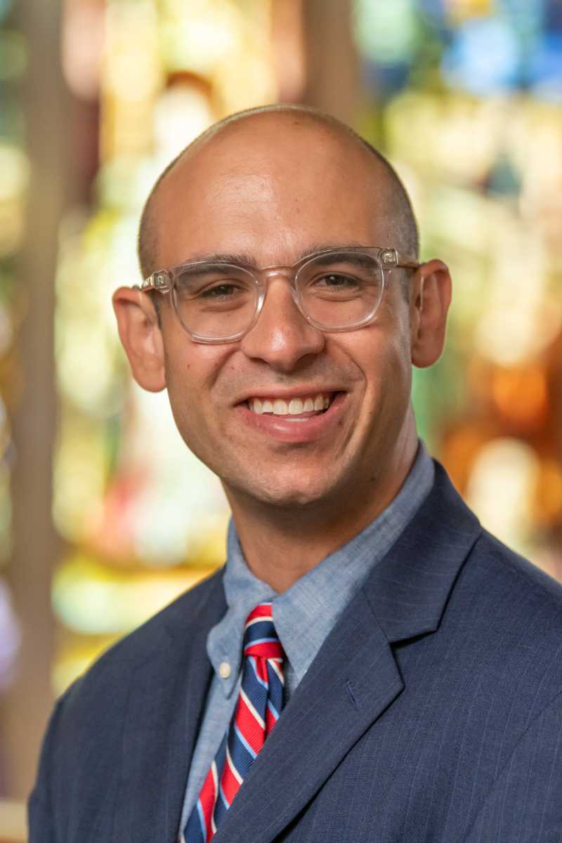 A portrait of Pasquale Toscano, a person with no hair, glasses, and a dark blue suit jacket.