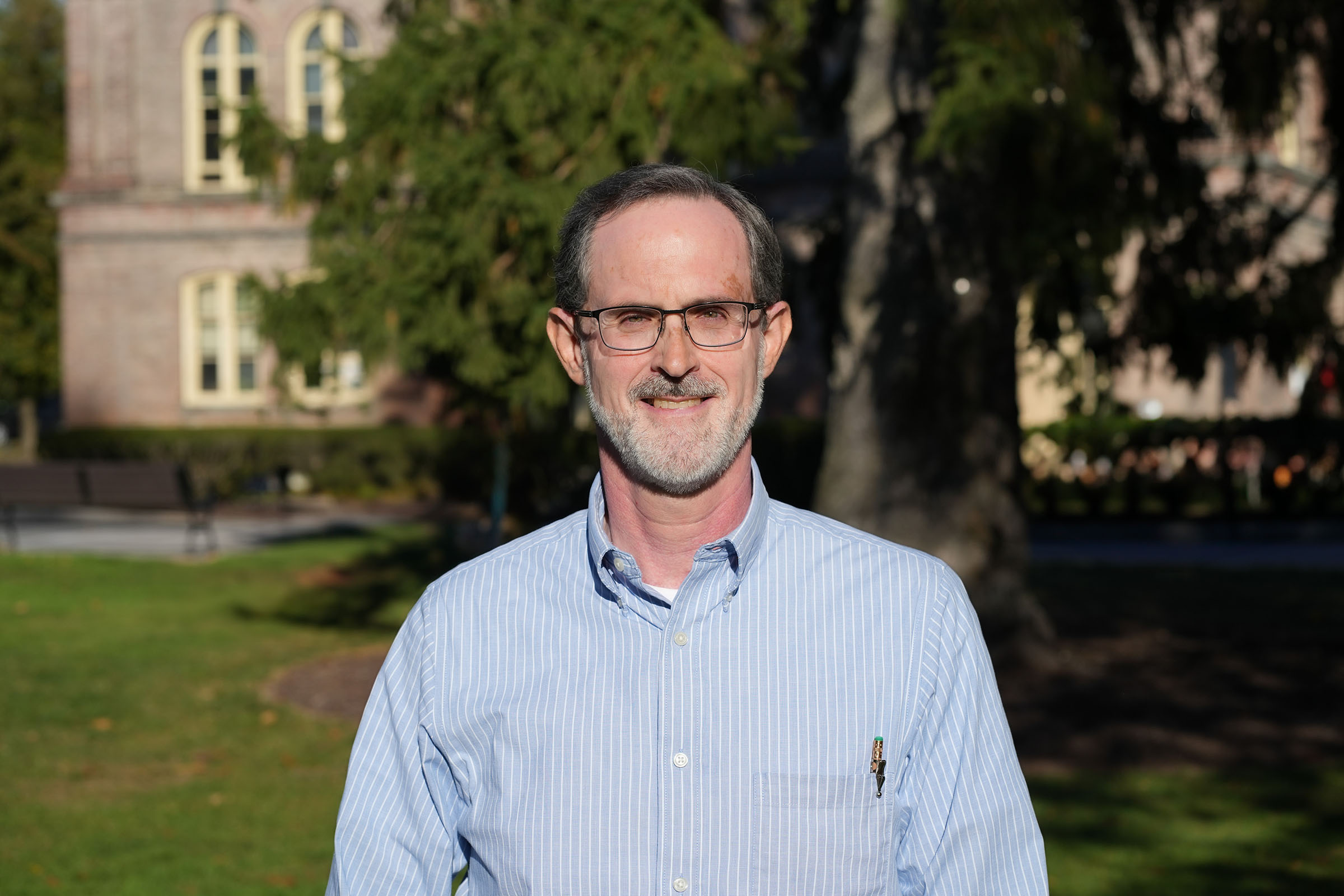 Ken Foster wearing a light blue with white striped collared shirt, eyeglasses, and short hair with beard and mustache with a brick building and large tree in the background.