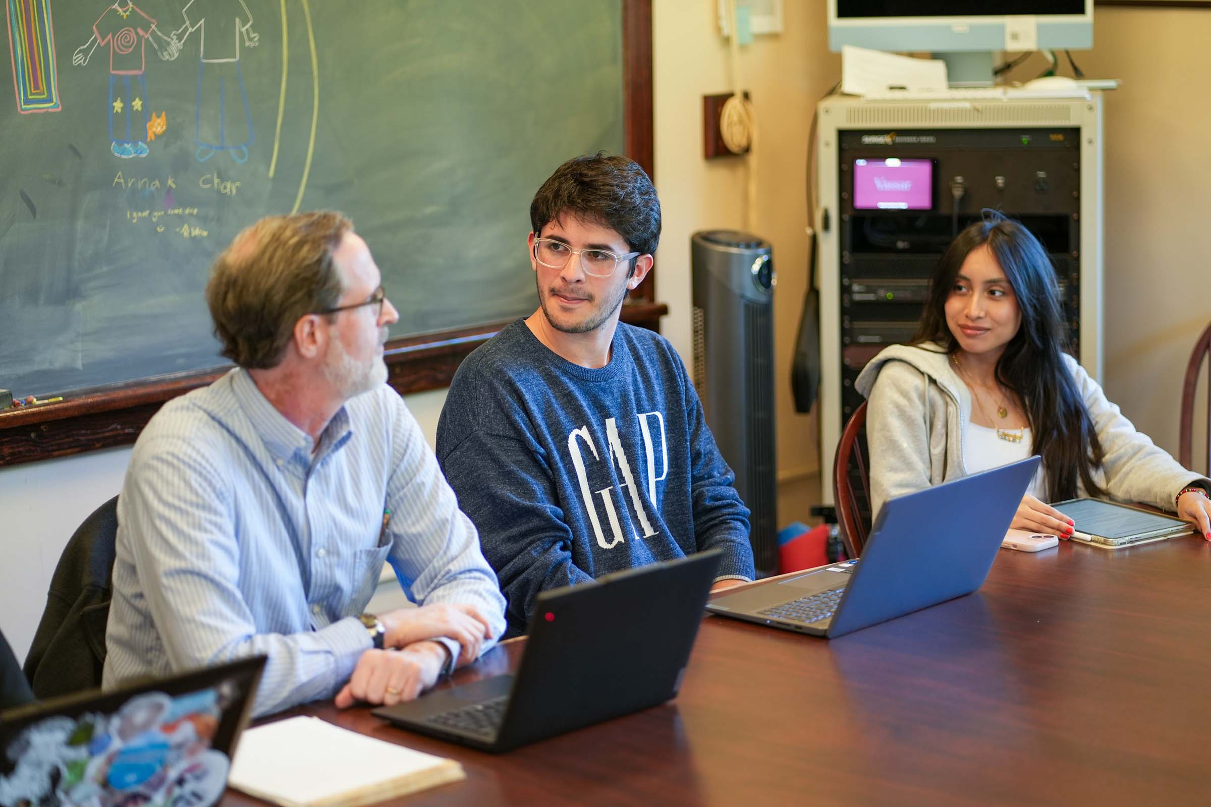 Three people sitting at a table in front of laptops looking at each other with chalkboard in the background.