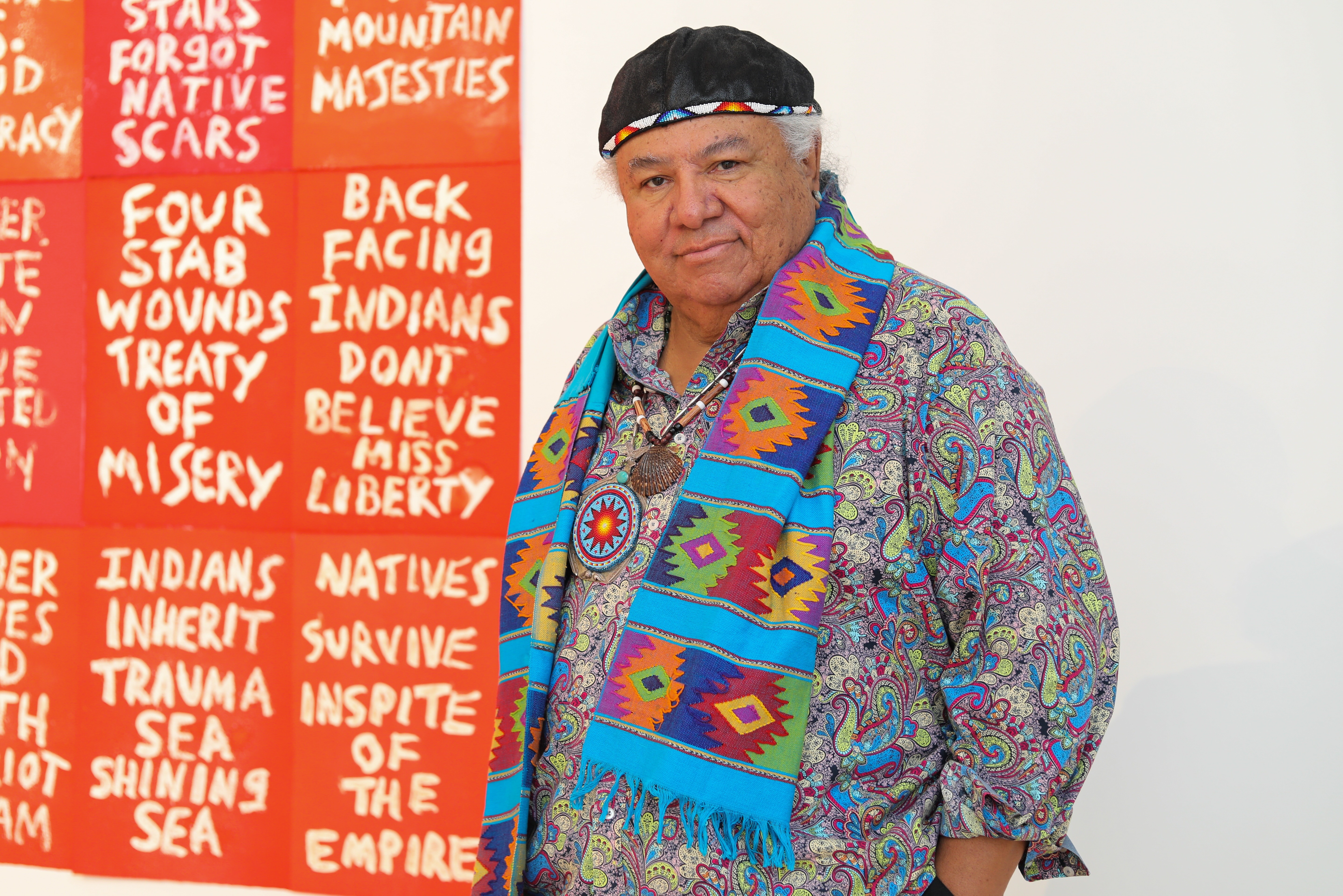 Photo of artist Edgar Heap of Birds standing in front of one of his text-based works. 