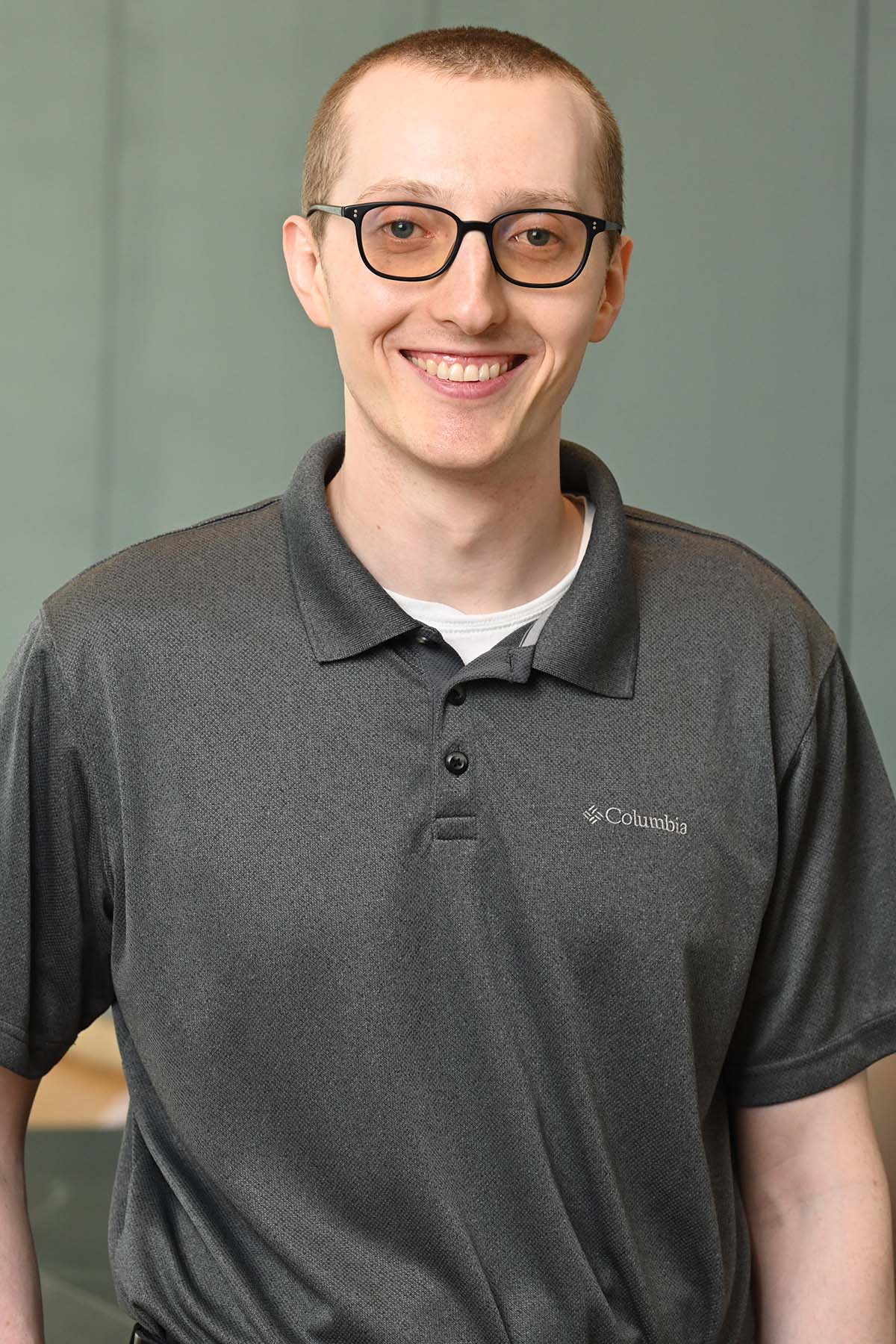 Person wearing a gray collared shirt over a white shirt, black framed eyeglasses, and short light hair. 