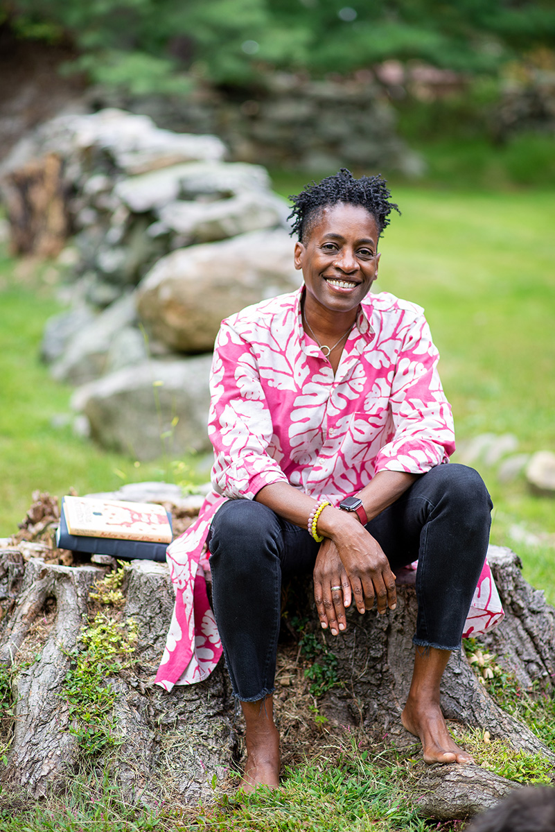 Person with dreadlocks and bright pink flowered shirt sitting outside on a tree stump with a low rock wall in the background with jeans and bare feet.