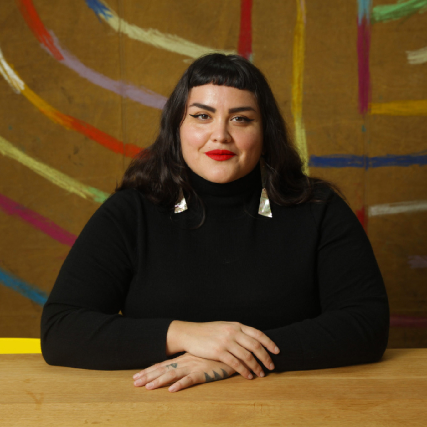 Pictured: Sarah Biscarra Dilley. A person sitting at a table with their arms crossed. The person is wearing a black shirt and long black hair with long earings.