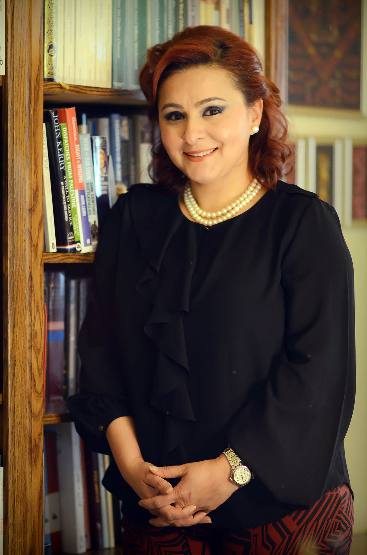 Shahla Ujayli wearing a black long sleeved shirt, red and black pants, and double strand of pearls with bookshelves in the background.