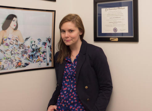 Dr. Andrea McDonnell, a person with long brown hair and a black coat, stands in front of a wall with diplomas and a painting hung on it.