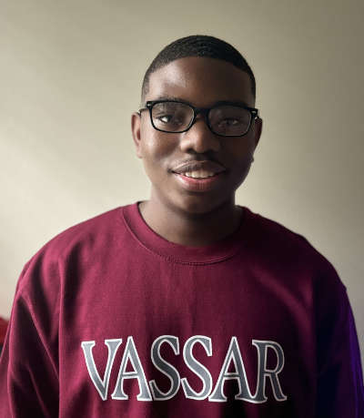 A portrait of Aryon Turner ’28, a person with glasses, short dark hair, and a maroon shirt with "Vassar" on it.