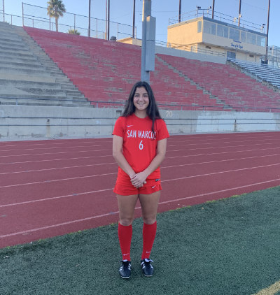 A portrait of Becca Rodriguez ’26, a person with long brown hair. Becca is wearing a red sports uniform with "San Marcos 6" on it in white lettering.