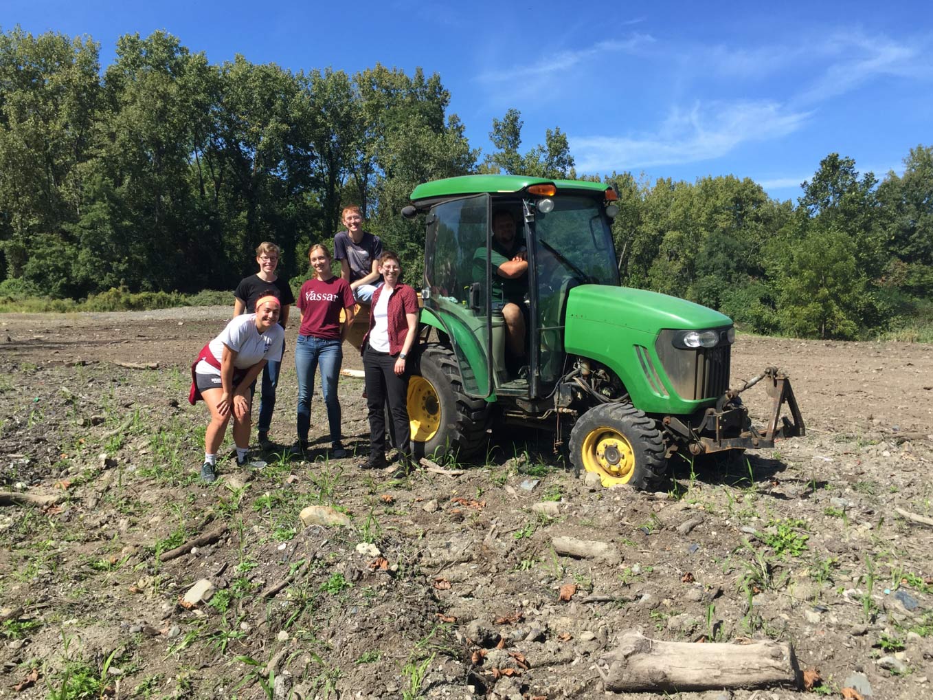 A group of students pose around a green tractor in a tilled field.