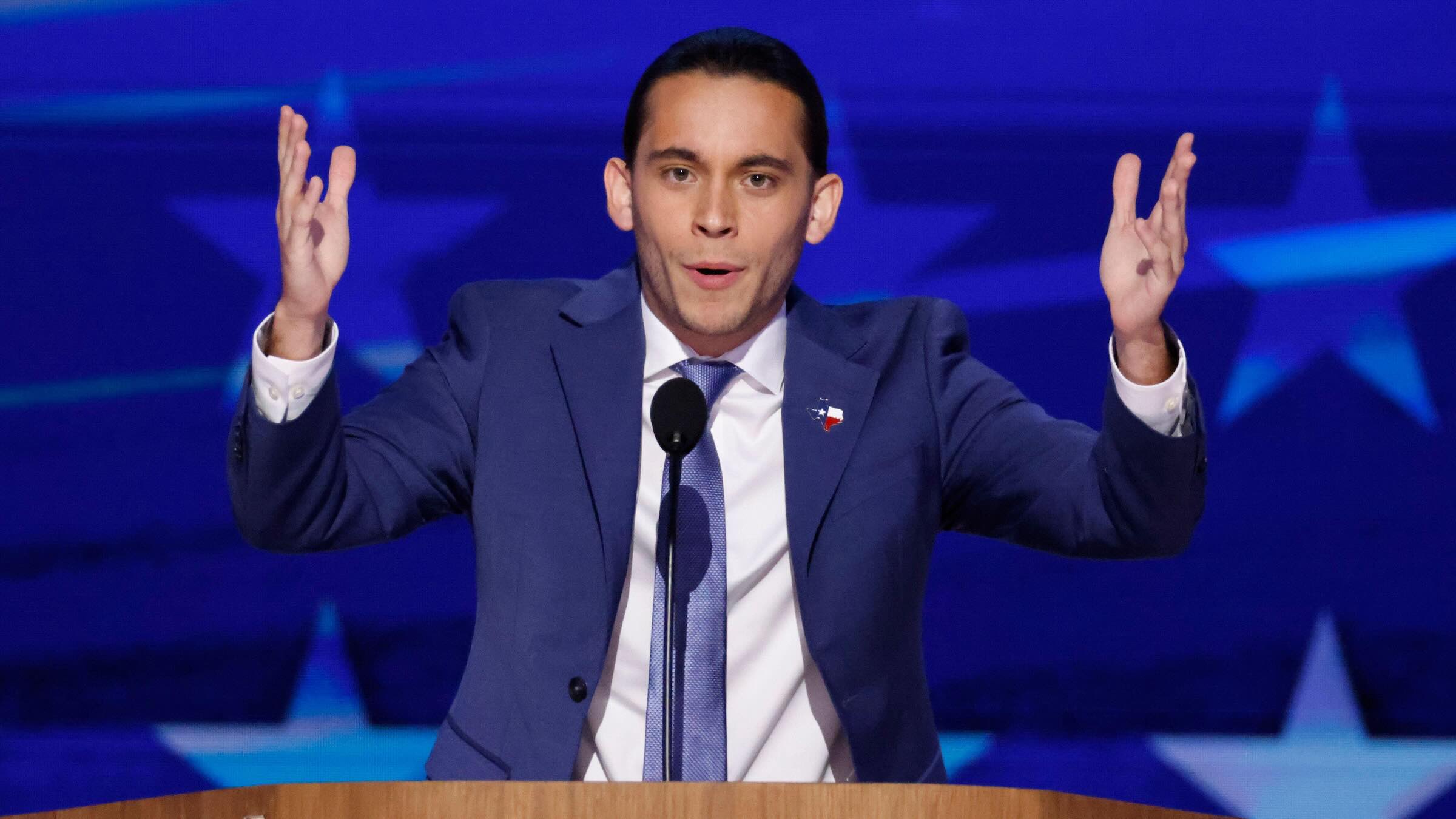 Person standing behind and lectern and microphone with both arms in the air speaking emphatically.