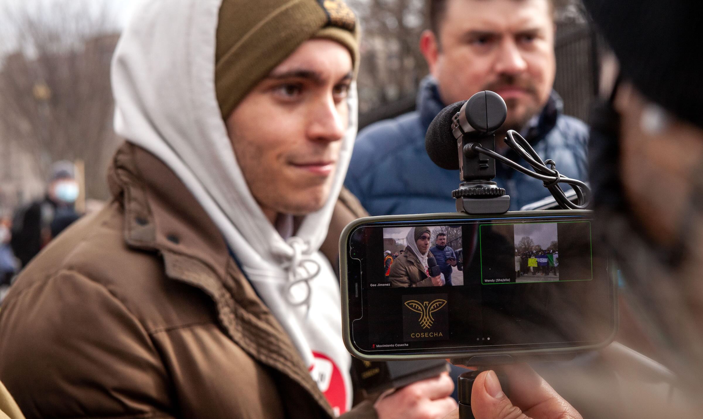 Person holding a microphone and dressed for the cold being filmed on a phone/tablet in the forground.