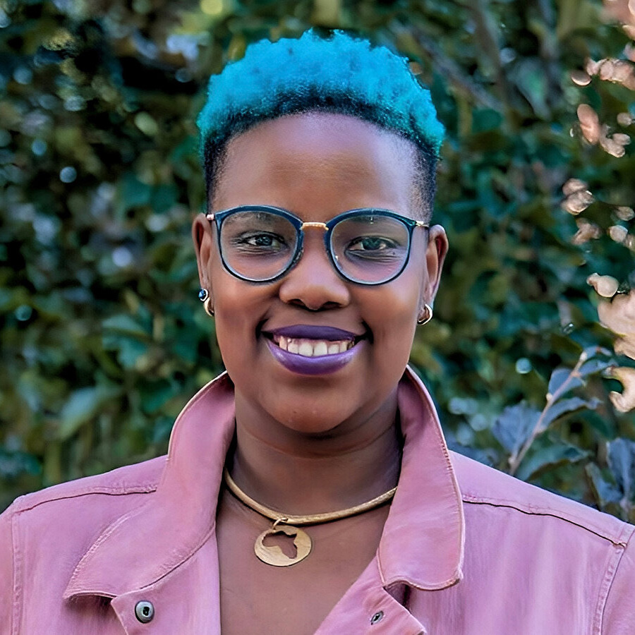 Very color image of a person with vivid, close-cut, light-blue hair. The person is wearing glasses, gold necklace, and pink oxford collared shirt while standing outside.