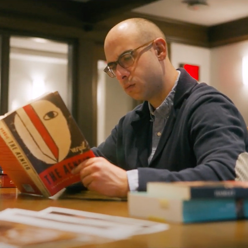 Person with glasses sitting at a table in a library holding up a book while reading it.