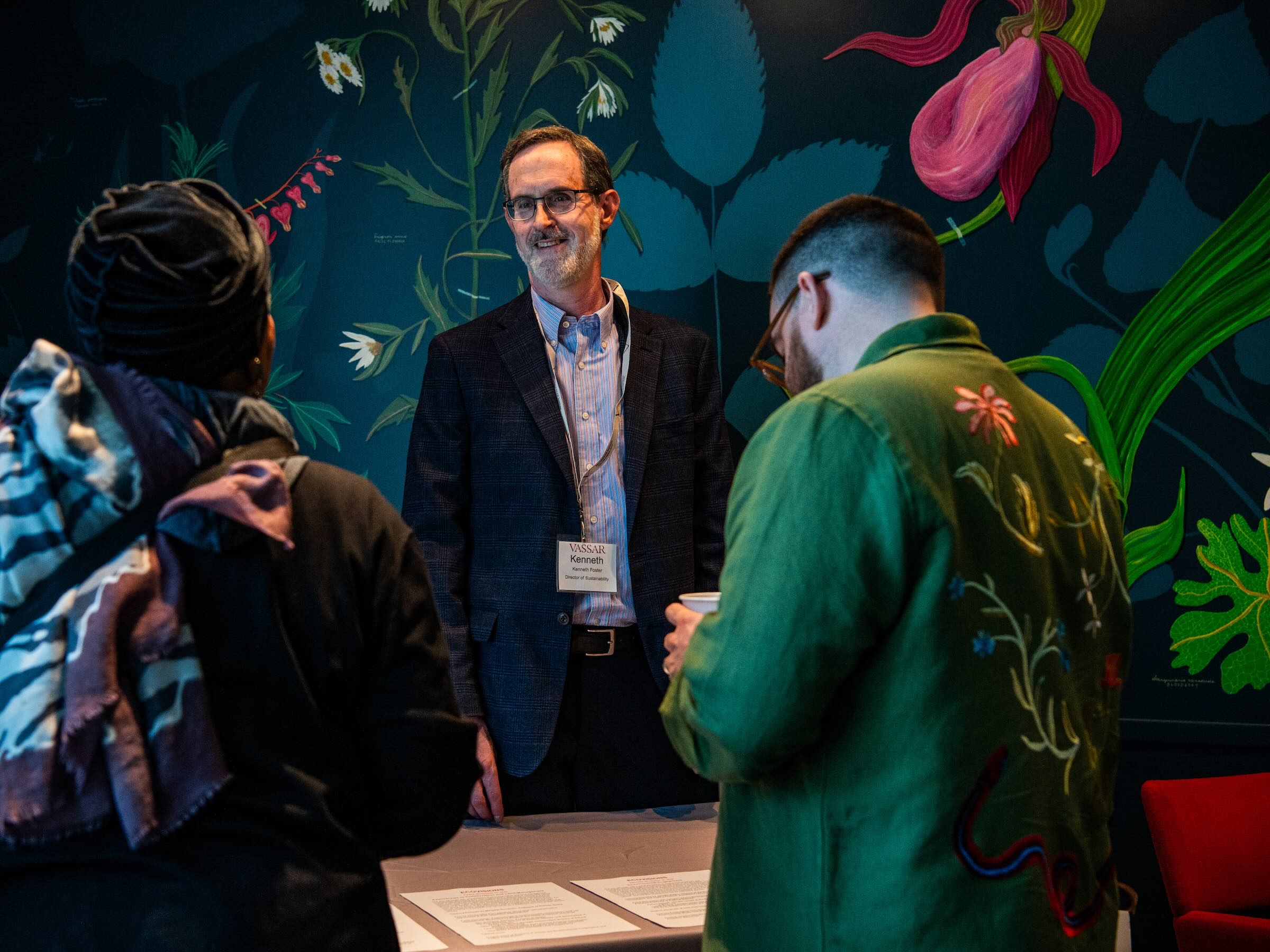 A smiling person standing behind a table answering questions for people standing in front of the table.