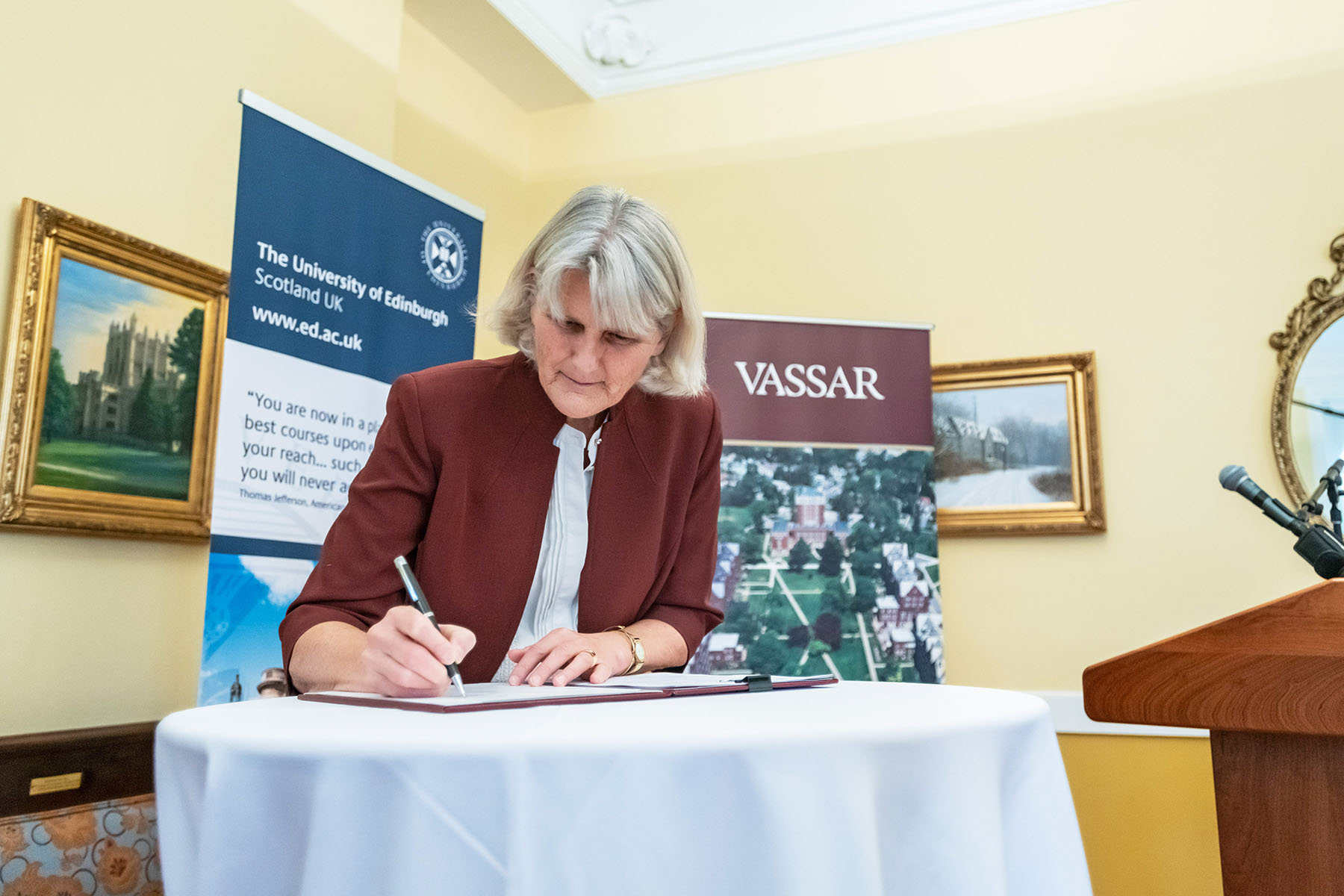 A person signing a document on a round linen covered table in a room with pictured adorning the walls.