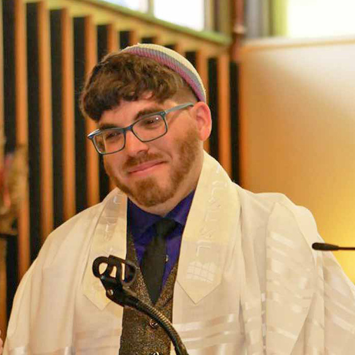 Rabbi Bryan Mann at a microphone wearing a purple shirt, black tie, gray vest, white robes, yarmulke and eyeglasses.