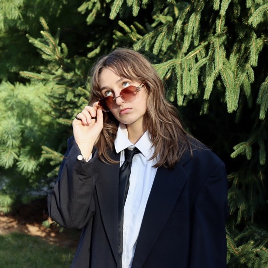 A portrait of Bella McCray, a person with long brown hair, sunglasses, a black coat, and a white shirt.