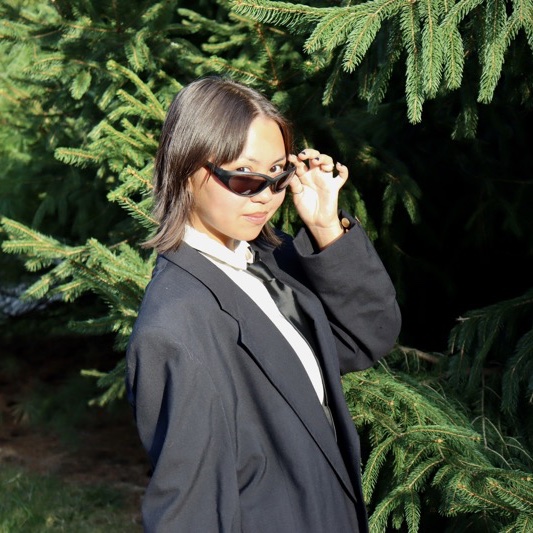 A portrait of Dana Tagliaferro, a person with long dark brown hair, sunglasses, and a black coat.