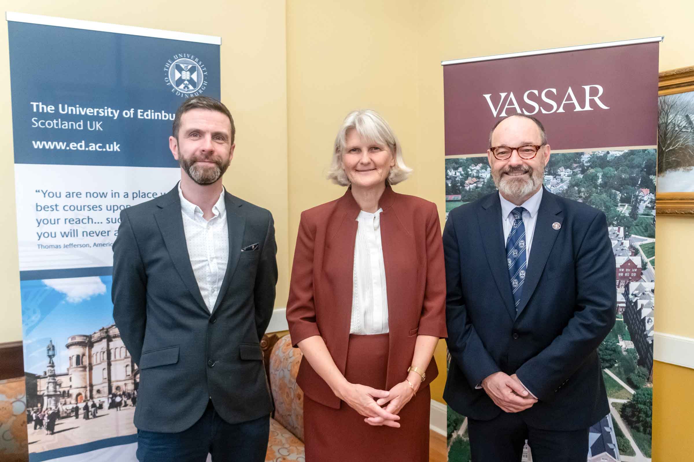 Three people standing and smiling in front of banners that read: University of Edinburgh and Vassar.