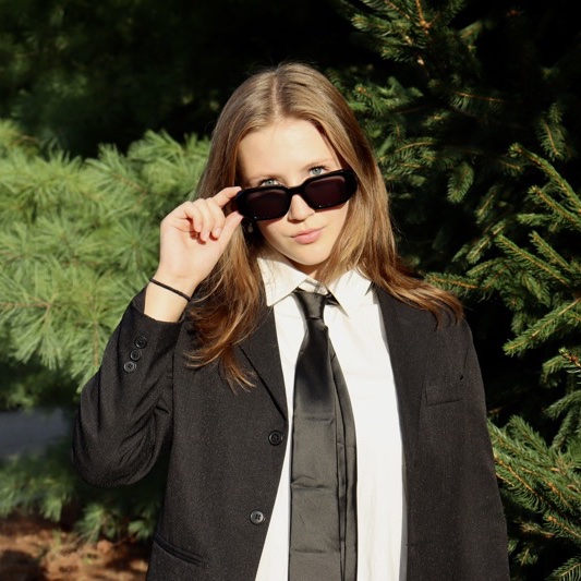 A portrait of Ella Albrecht, a person with long brown hair, sunglasses, a black coat, and a white shirt.