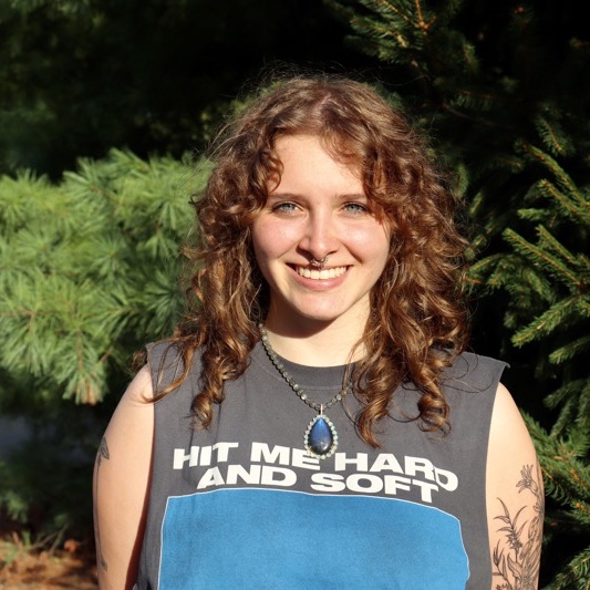 A portrait of Frieda Dangerfield, a person with long curly red hair and a gray shirt that says "Hit me hard and soft."