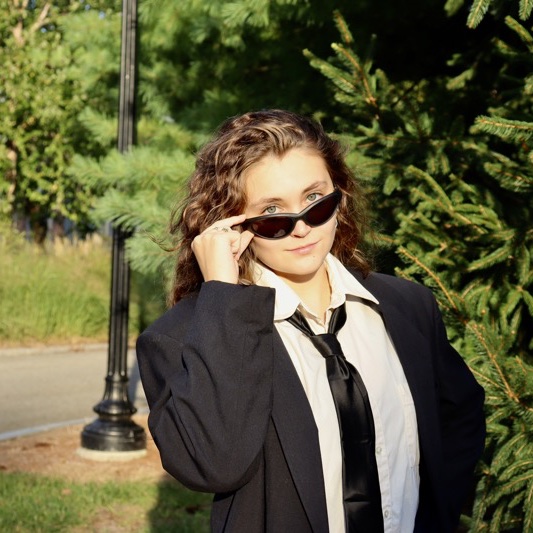 A portrait of Helen Ambrose, a person with long brown hair, sunglasses, a black coat, and a white shirt.