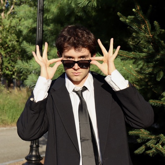 A portrait of Jacob Geiger, a person with short brown hair, sunglasses, a black coat, and a white shirt.