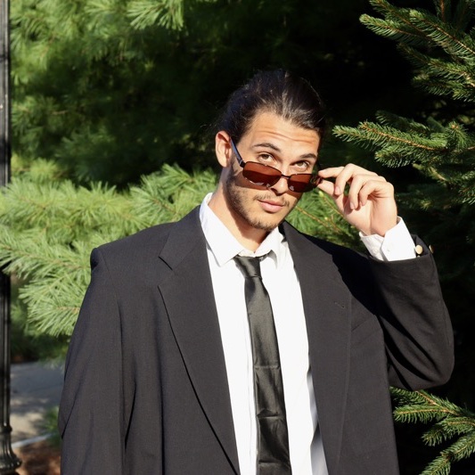 A portrait of Ki’tsai Zangpo, a person with short dark brown hair, sunglasses, a black coat, and a white shirt.