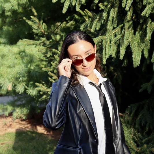 A portrait of Maia Beaudry, a person with long black hair, sunglasses, a black coat, and a white shirt.