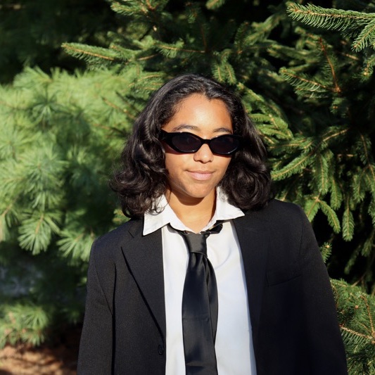 A portrait of Nia Bethel-Brescia, a person with long dark brown hair, sunglasses, a black coat, and a white shirt.